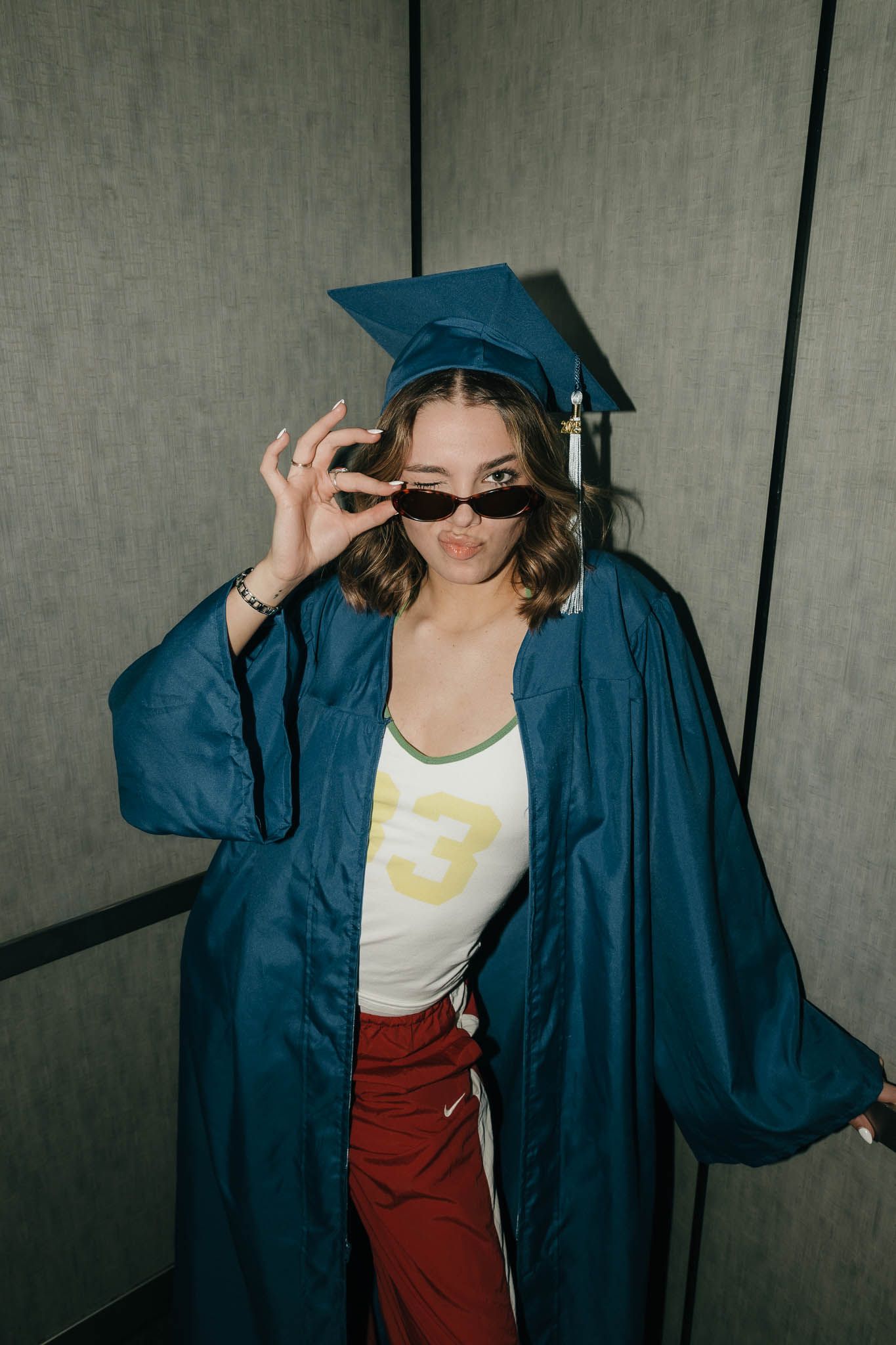 Graduate in blue cap and gown wearing sunglasses and red pants strikes a playful pose in hallway.