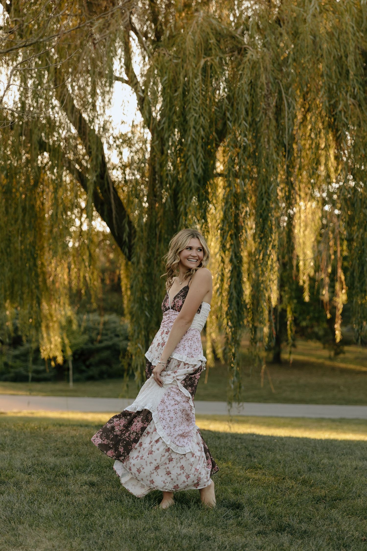 Woman in floral dress twirls gracefully under weeping willow trees at golden sunset in park setting.