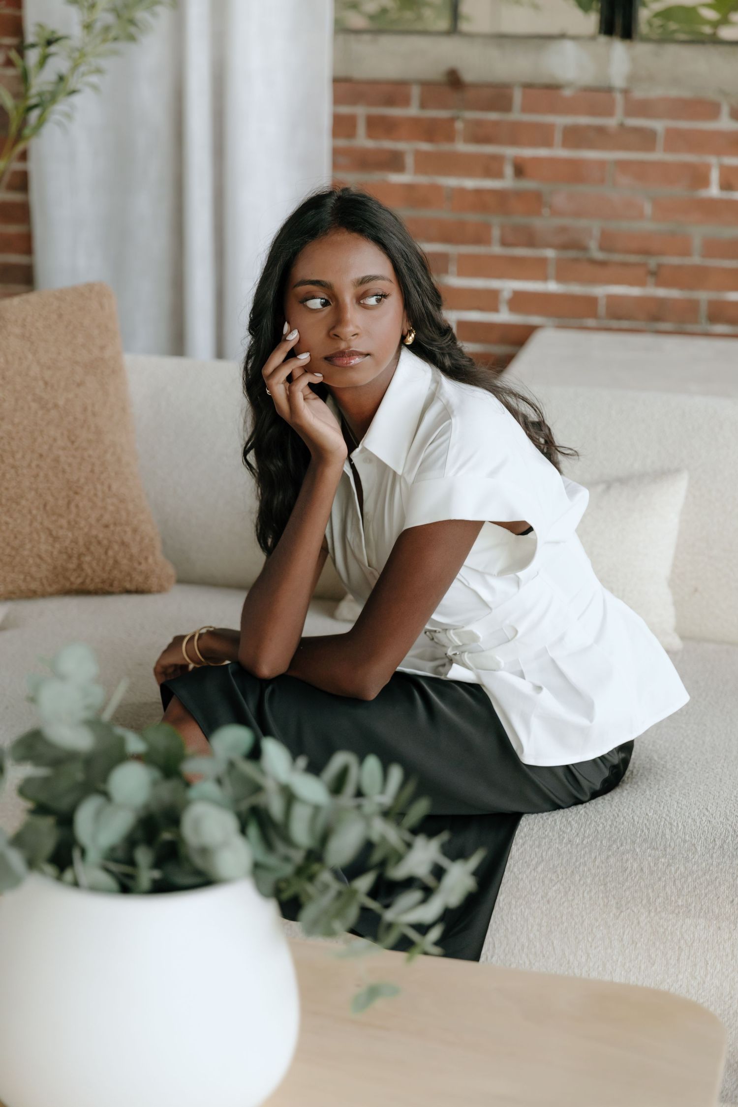 Young Black woman in white top and black pants sits on a cream sofa, posing thoughtfully with hand near face.