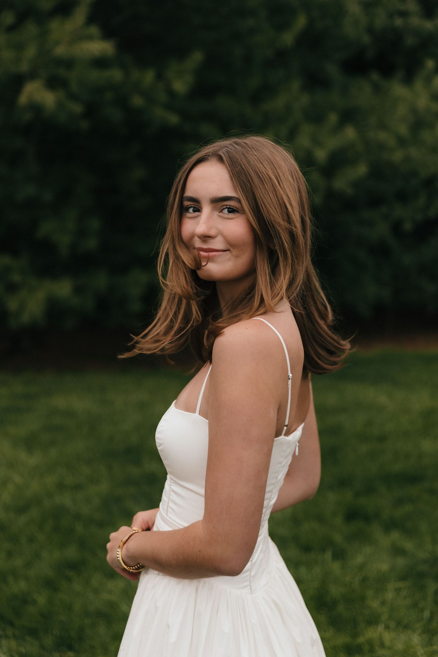 Young woman in white sundress with gold bracelet smiling over shoulder outdoors with lush green background.