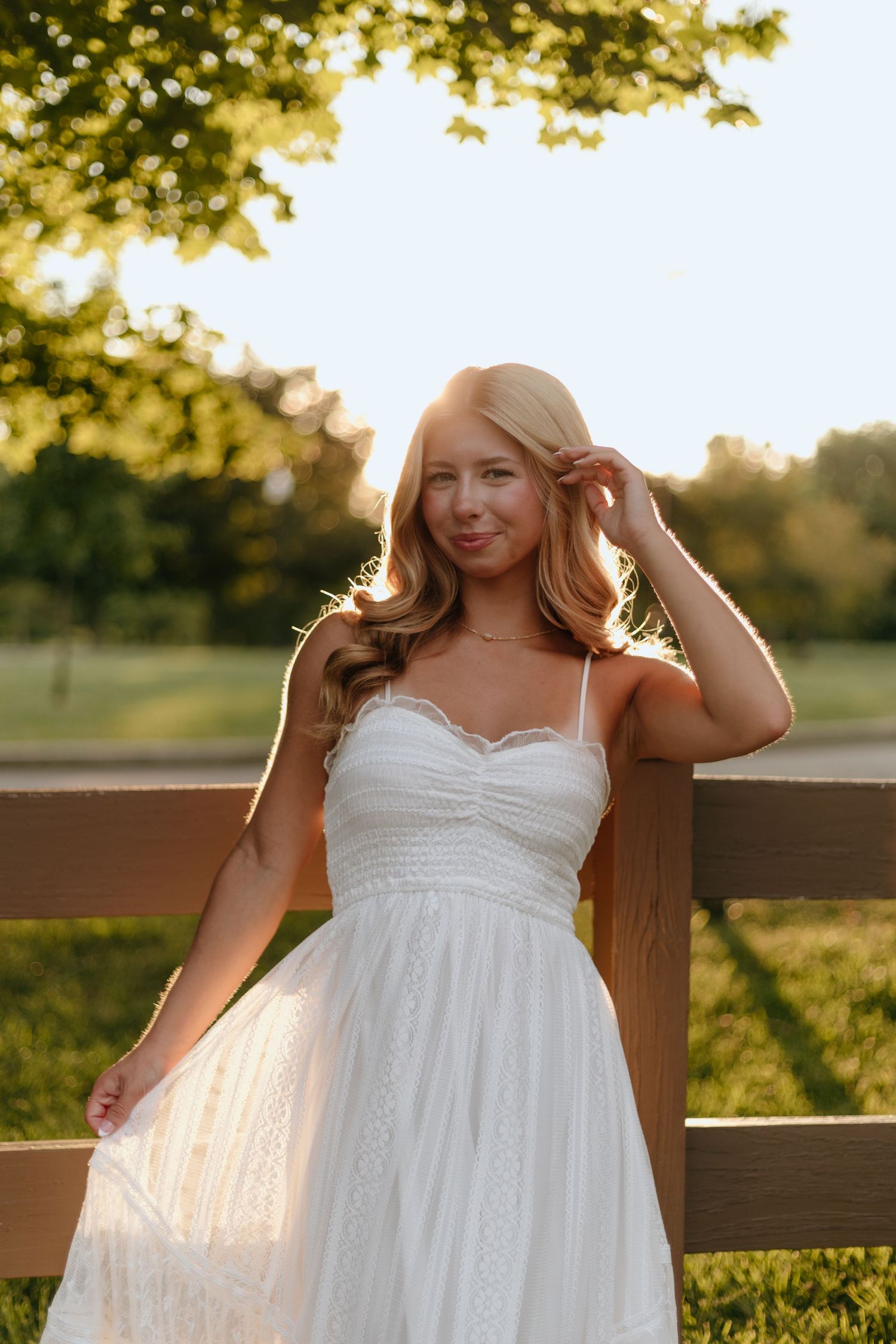 Woman in white sundress poses by wooden fence during golden hour.
