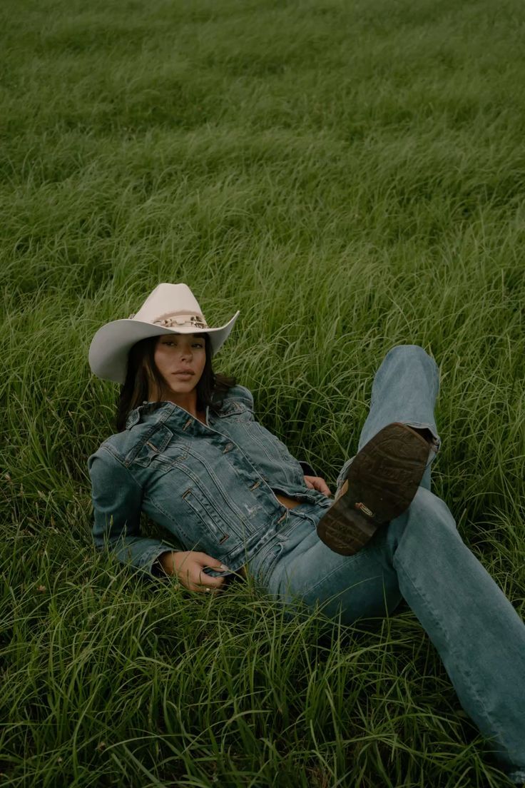 Person in denim and cowboy hat lies in tall green grass field on a cloudy day.