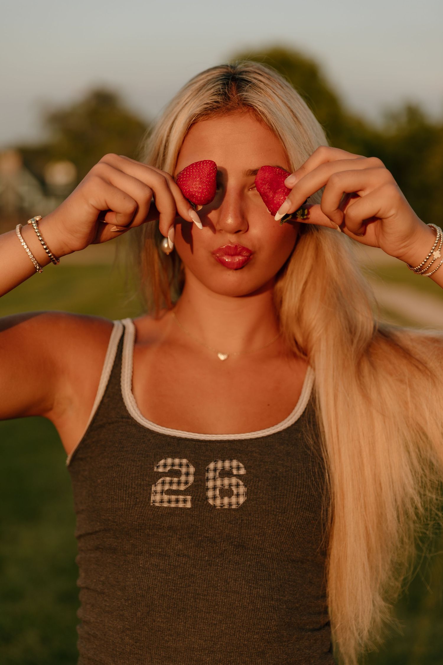Blonde woman in gray #26 tank top holding strawberry halves over her eyes, posing outdoors at golden hour.