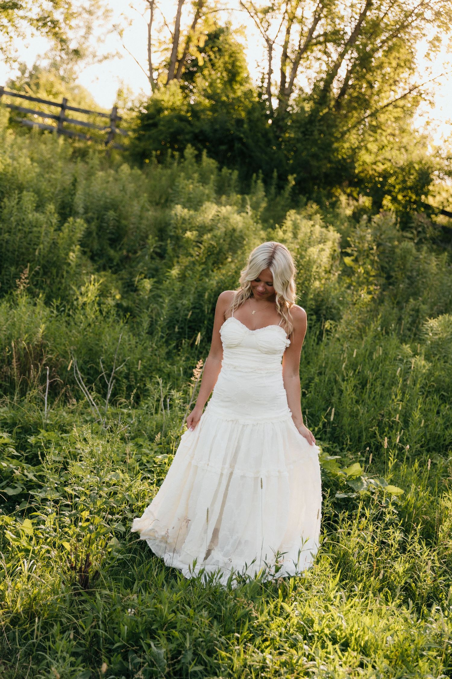Blonde bride in strapless white wedding gown standing in lush green meadow during golden hour sunset.