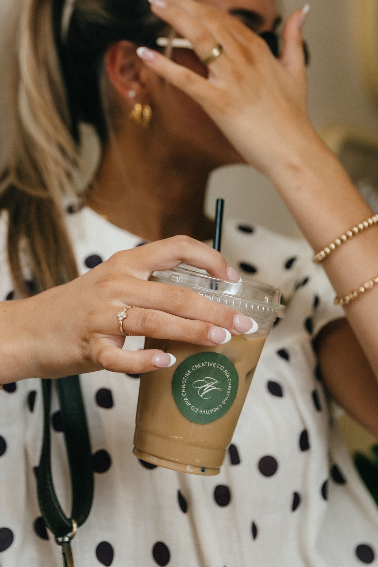Woman holding an iced coffee cup with a green logo, wearing gold rings and a polka dot top.