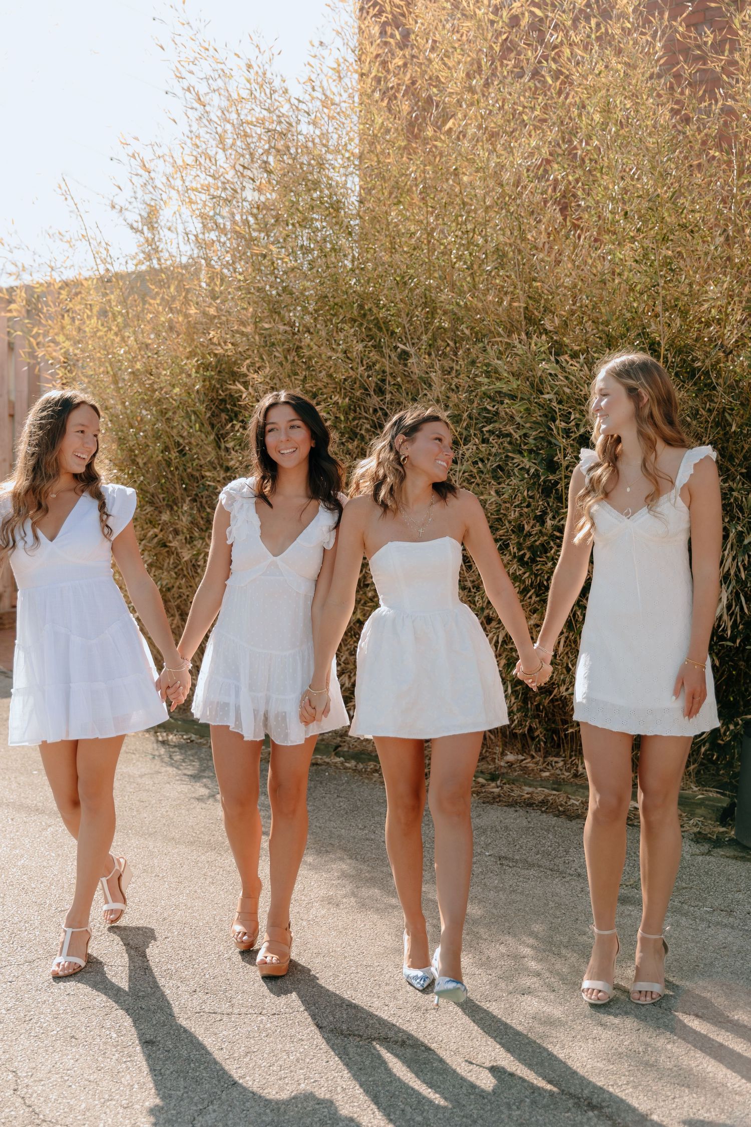 Four friends in white summer dresses walk together holding hands on a sunny path with golden foliage in background.