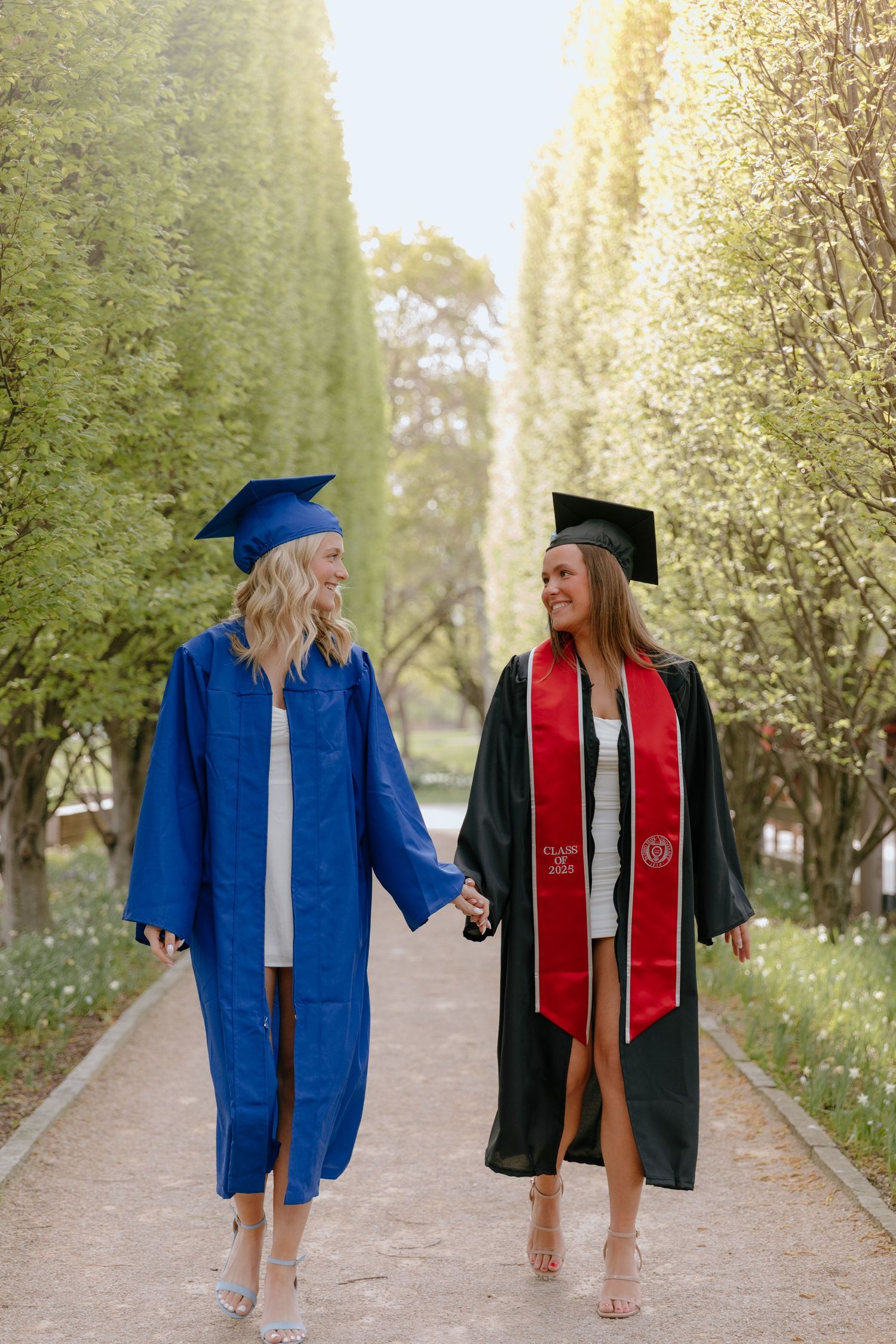 Two graduates in blue and red commencement gowns walking together on a tree-lined path during a sunny spring day.