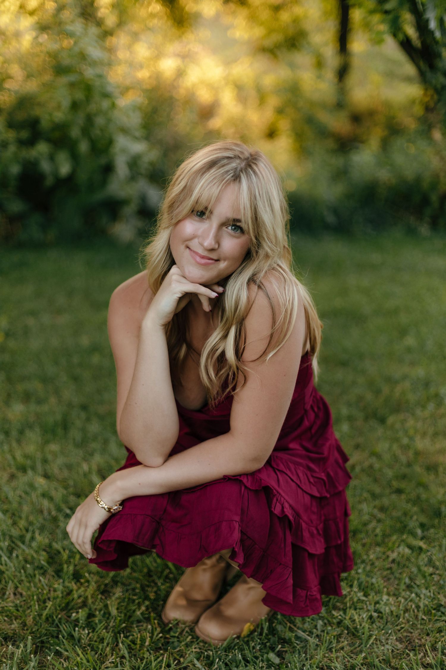 Person in burgundy dress poses gracefully on grass during golden hour outdoor photo session.
