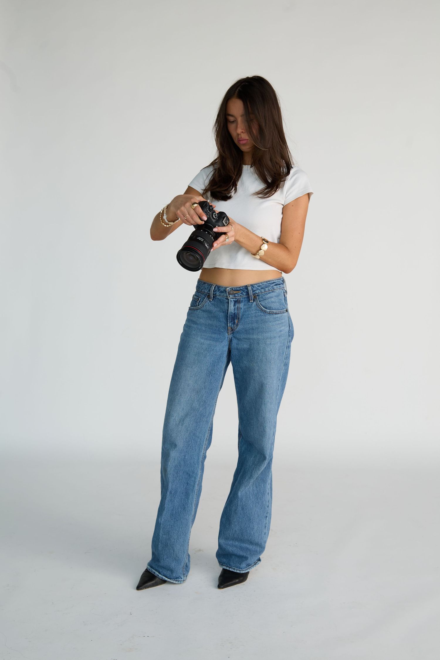 Young woman in white crop top and blue wide-leg jeans holding a camera, posing against a white background.