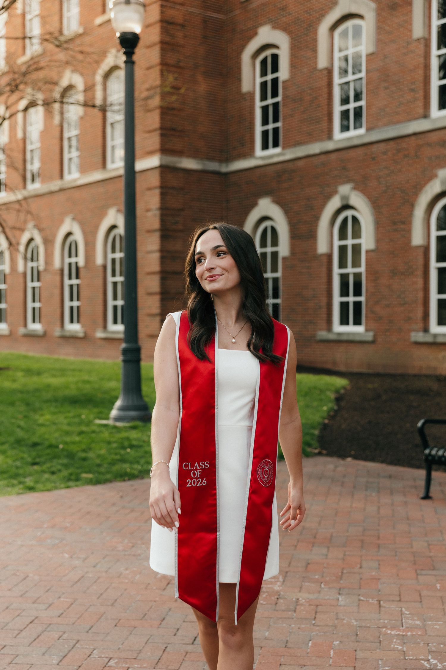 Young woman in white dress and red Class of 2026 graduation stole posing outside a brick university building.