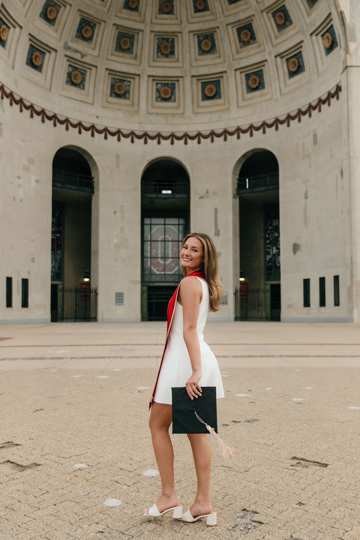 Smiling graduate in white dress holding diploma outside a grand domed building with ornate architecture.