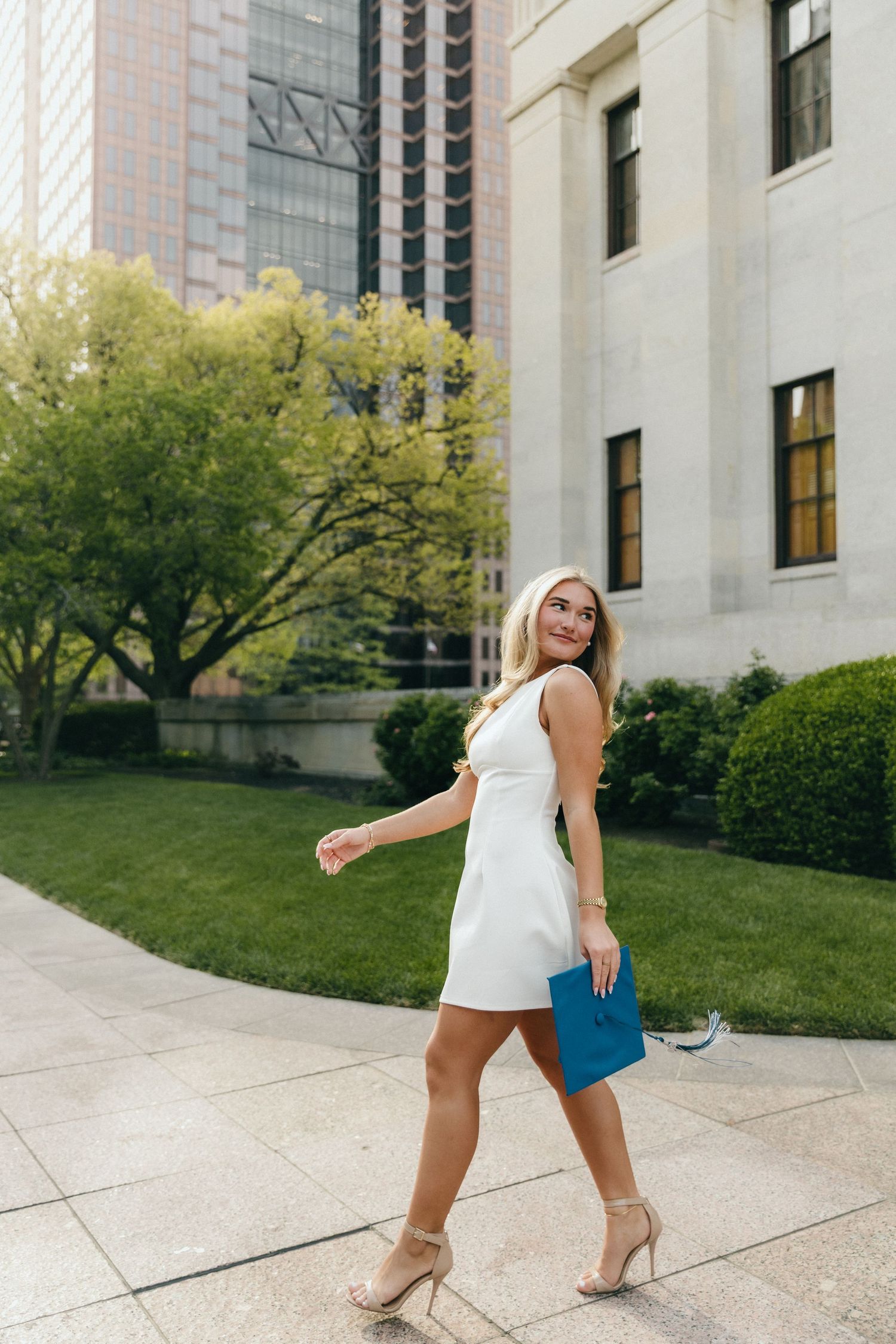 Young blonde woman in white mini dress and nude heels walks outdoors holding a blue clutch near city buildings.