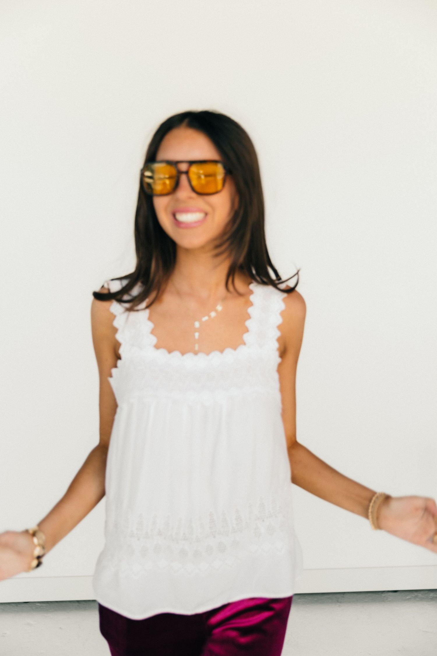 Smiling woman wearing orange tinted sunglasses, white ruffled tank top, and magenta pants against a white wall.
