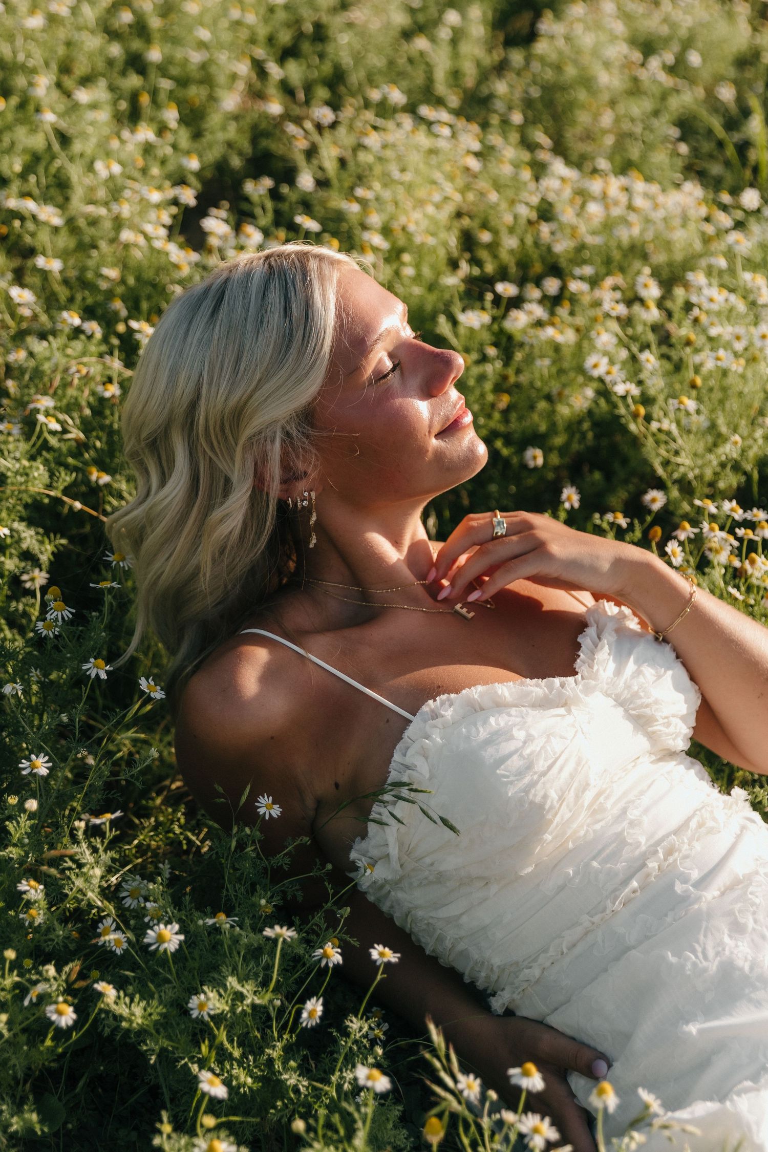 Blonde woman in white dress lying in a daisy field, eyes closed, basking in sunlight with elegant jewelry.