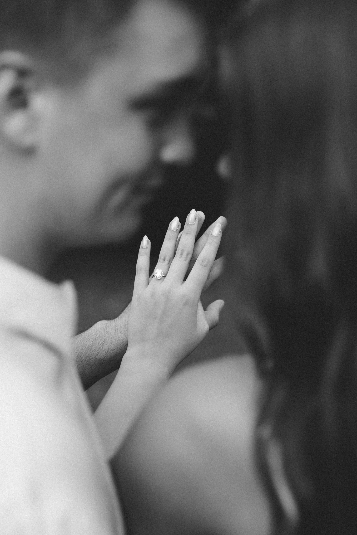 Black and white photo of engaged couple almost kissing while woman displays diamond engagement ring on her hand.