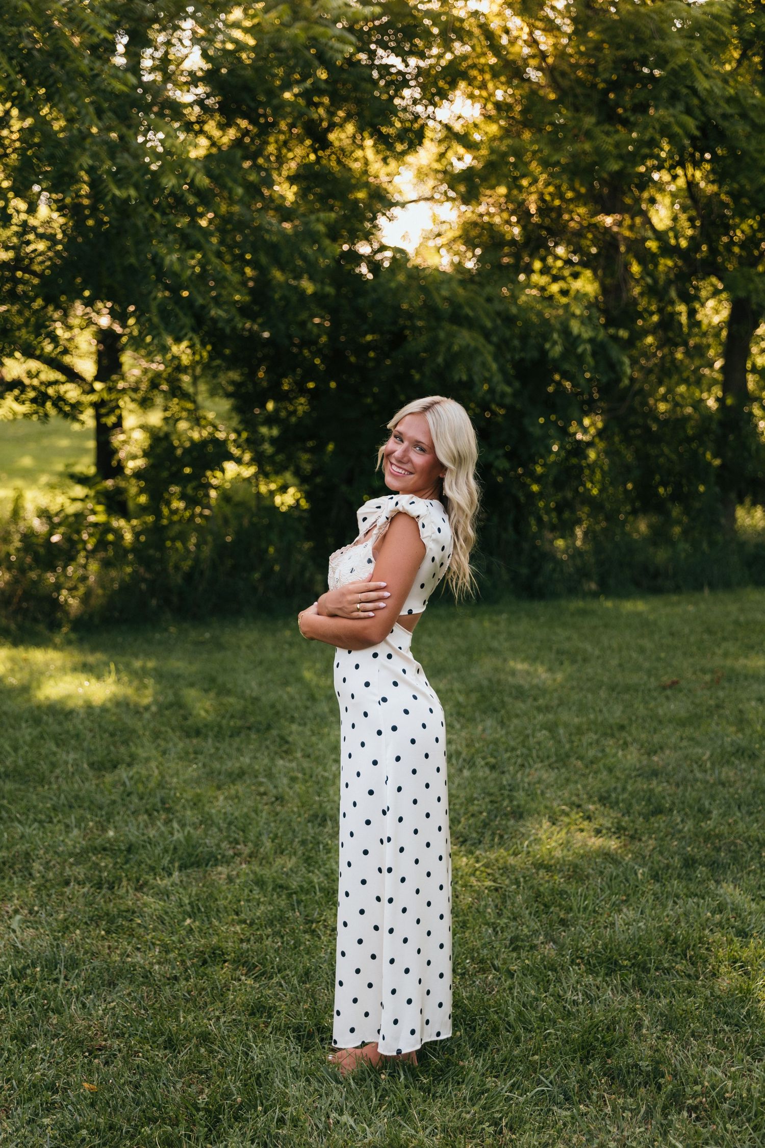 Smiling blonde woman in white polka dot dress standing on green grass with trees in background.