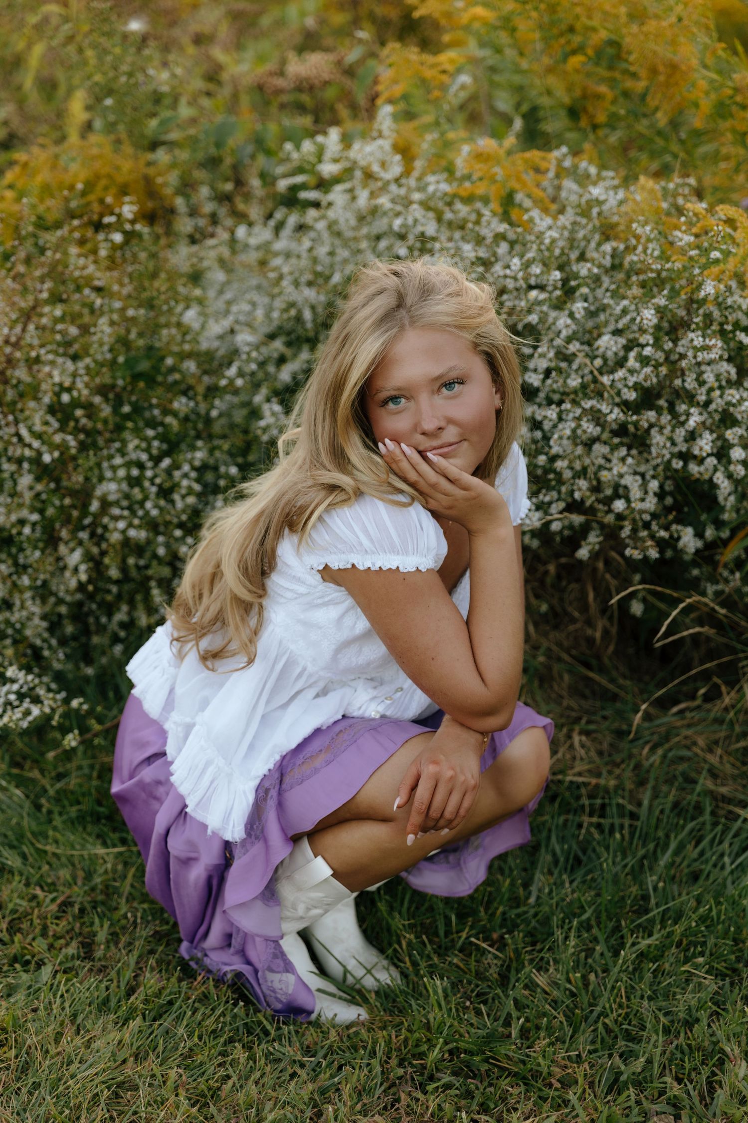 A child in a white and purple dress sits playfully in a garden with white wildflowers and green foliage.