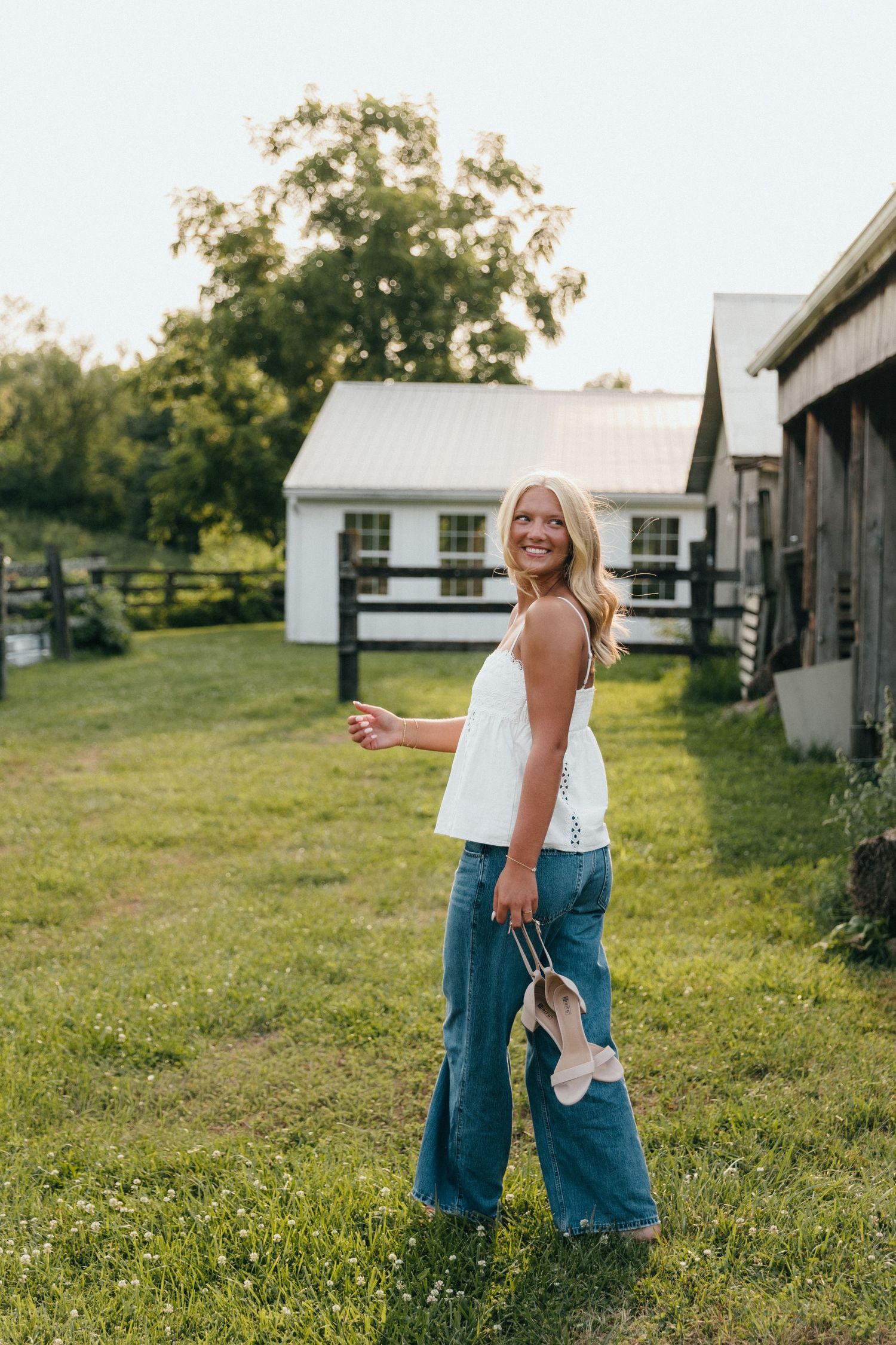 Smiling blonde woman in white top and wide-leg jeans holding shoes on a sunny farm.