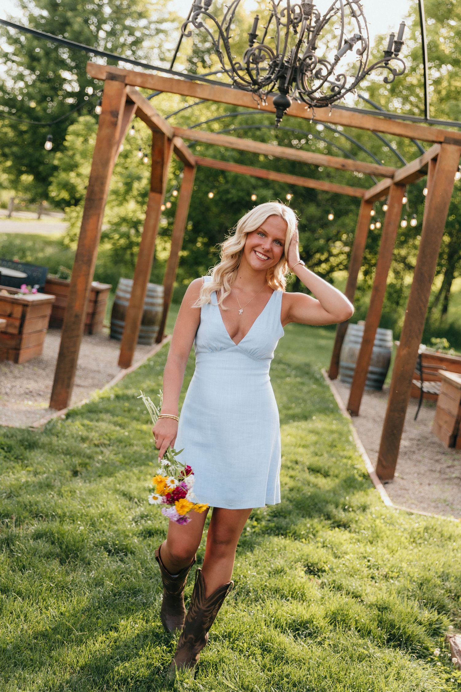Smiling blonde woman in light blue mini dress and cowboy boots holding wildflowers under a rustic wooden pergola.