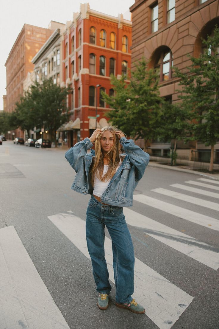 Person in oversized denim outfit playfully posing on a crosswalk with brick buildings in the background.
