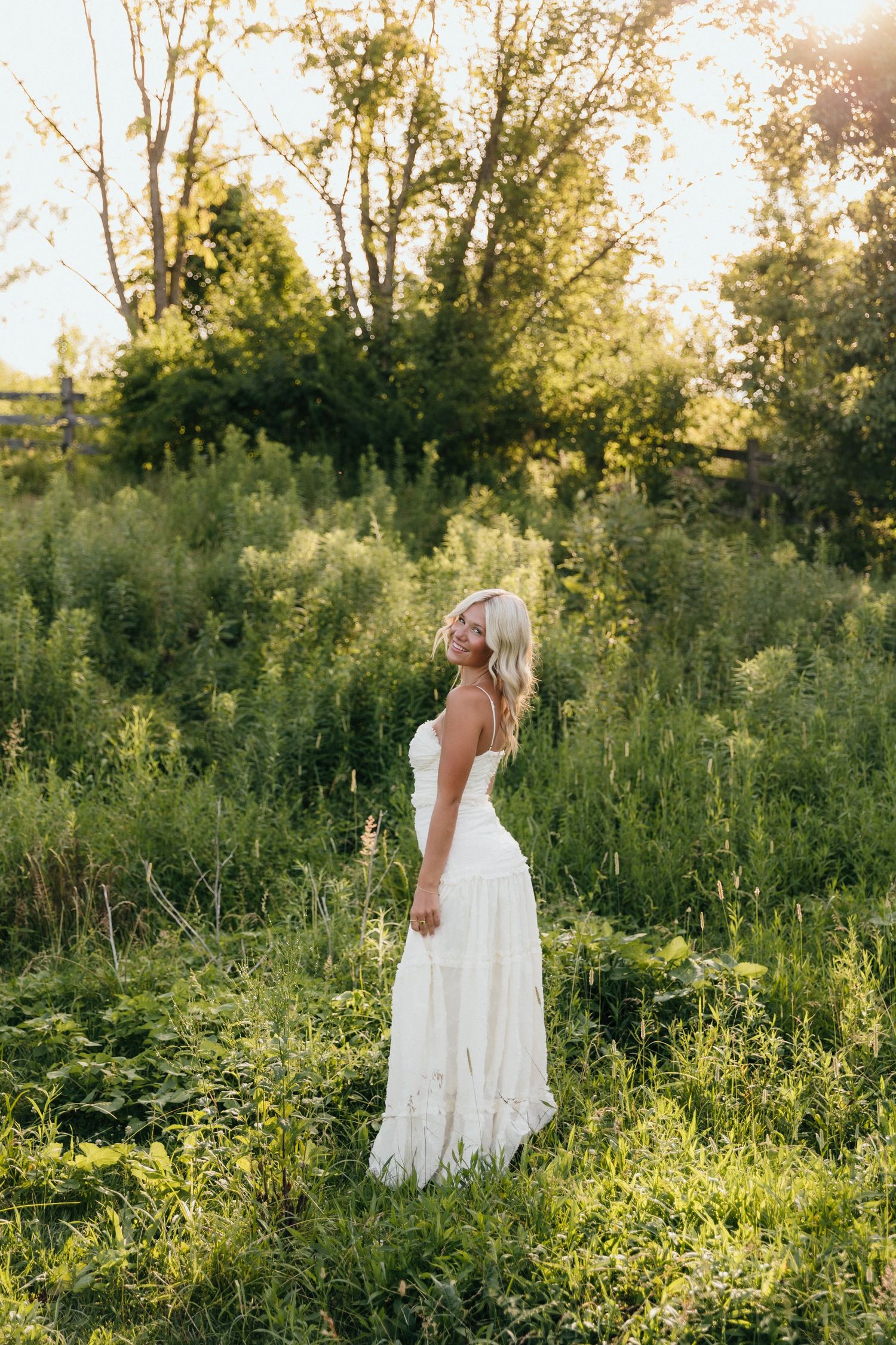 Blonde woman in white maxi dress standing in lush green field during golden hour sunset.