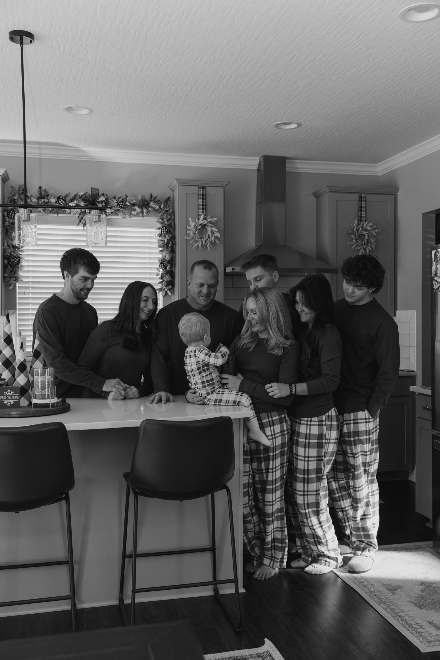 A group of people in black and plaid clothing gather around a kitchen counter in a black and white photograph.