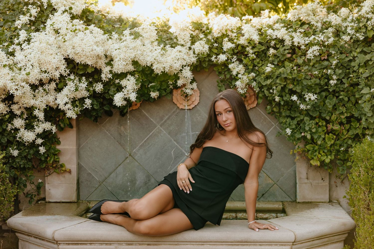 A figure in a black dress poses on a stone bench beneath a blooming white floral archway at sunset.