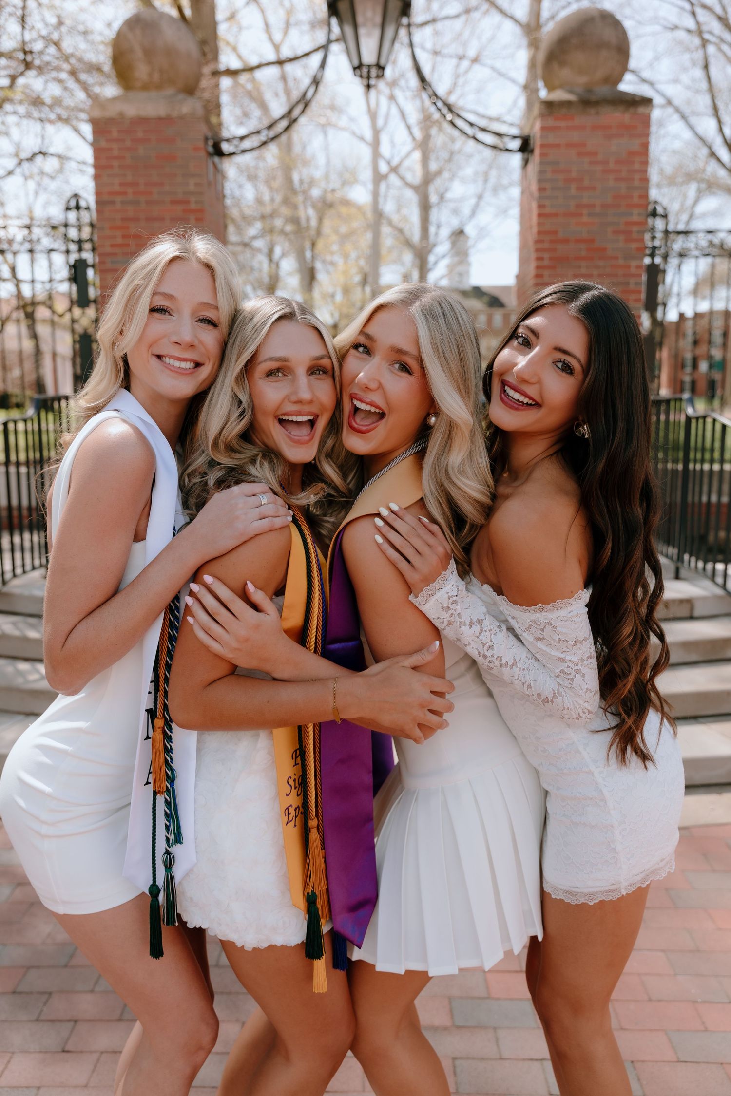 A group of friends in white and purple outfits laugh and embrace while posing together on a brick walkway.