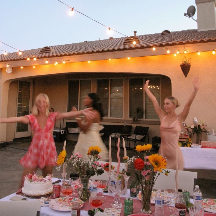 Three women dancing joyfully at an outdoor evening party with string lights, floral arrangements, and a decorated table.