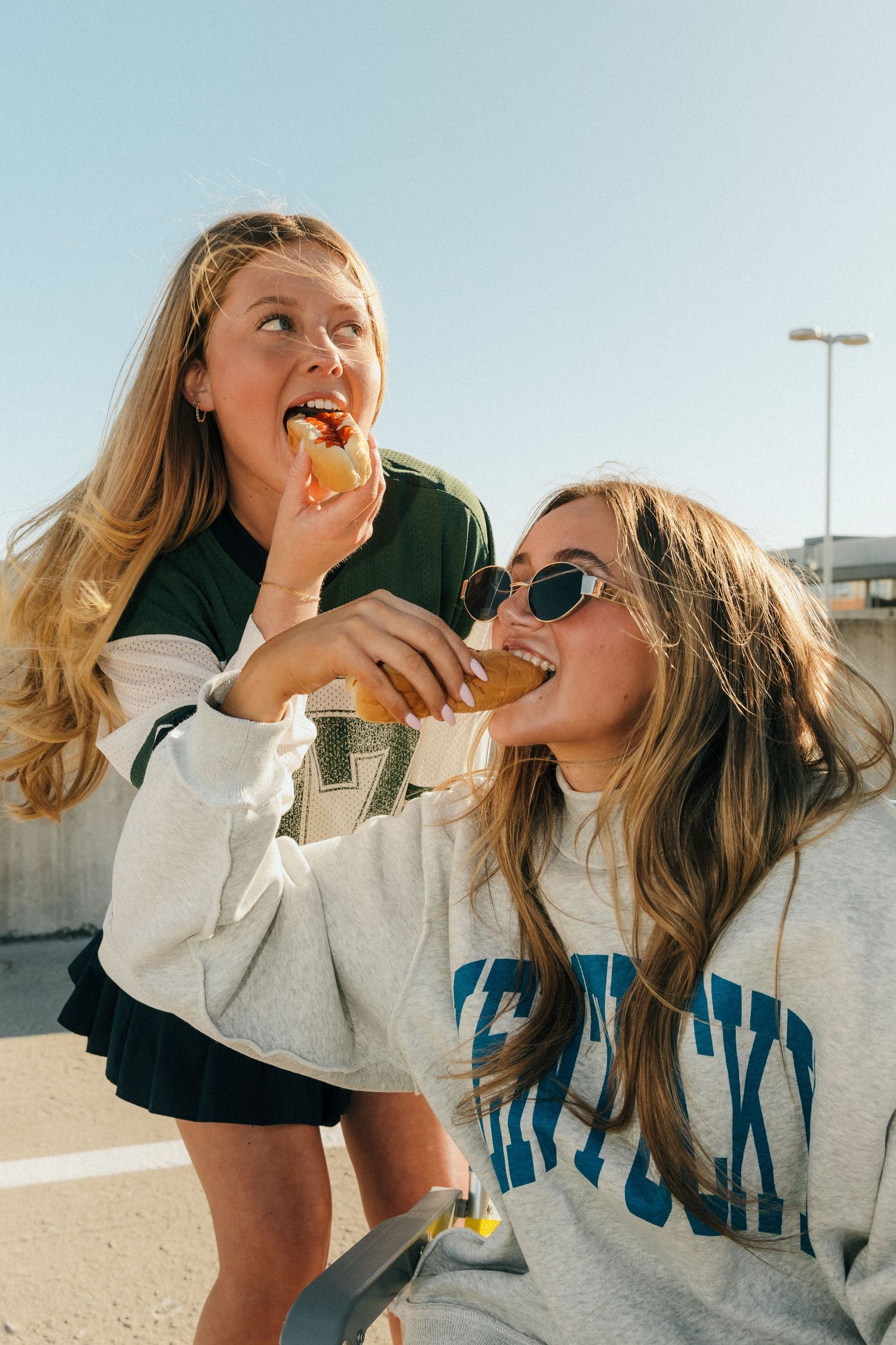 Two young women in college sweatshirts sharing snacks outdoors on a sunny day, smiling and enjoying food together.