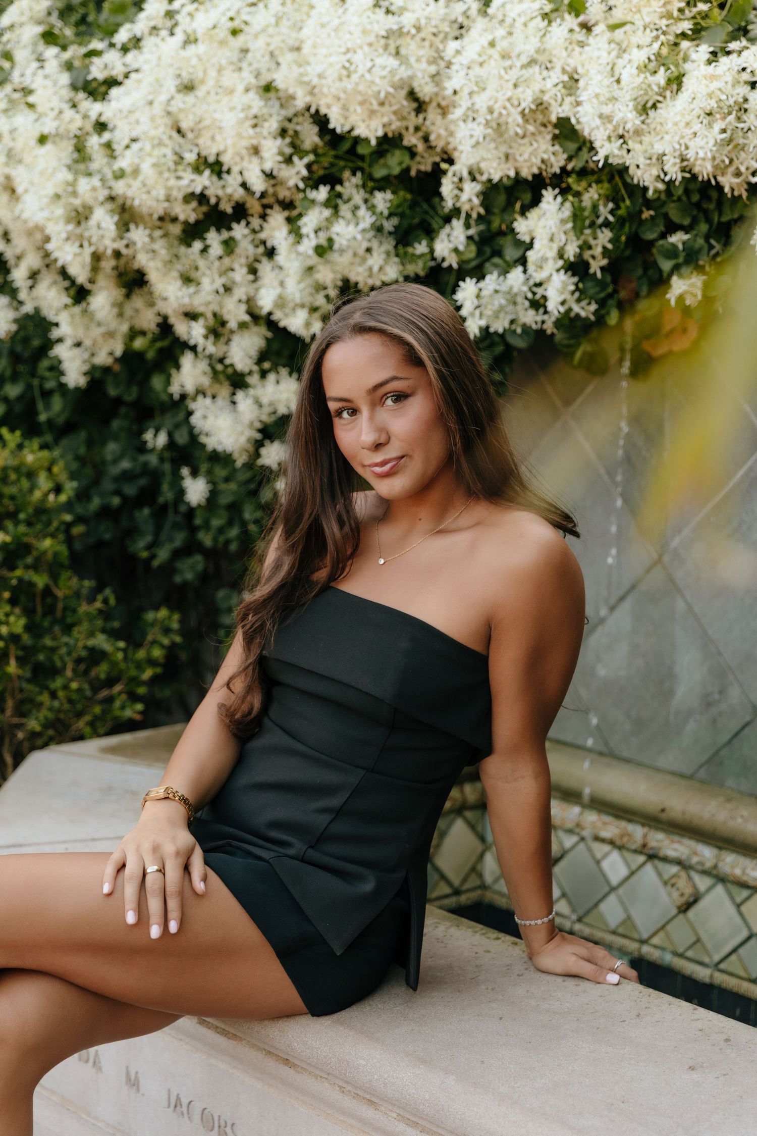 A woman in a black one-shoulder dress poses elegantly against white flowering branches in an outdoor setting.