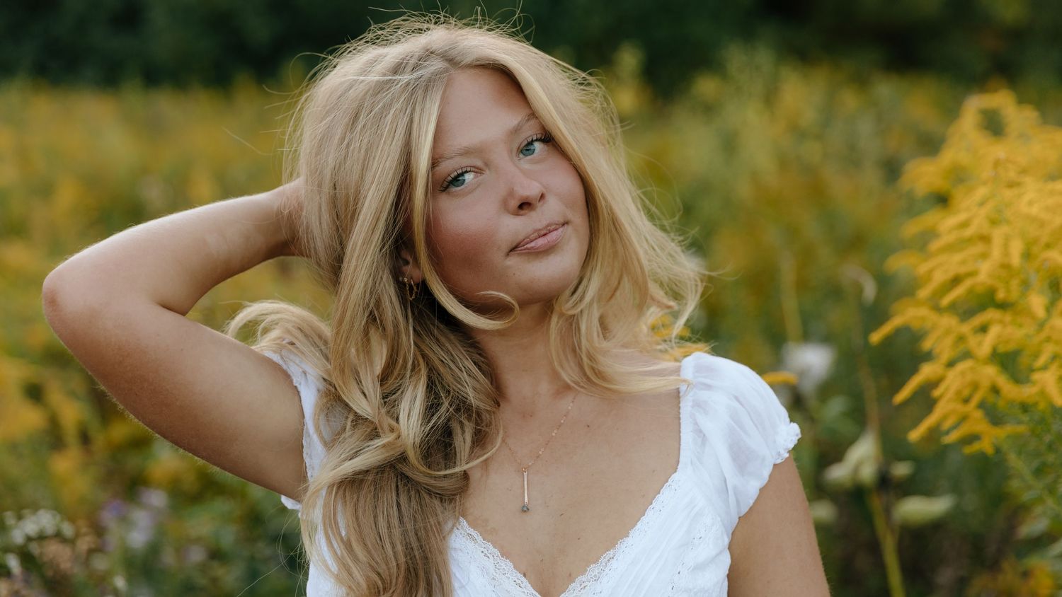 A person in a white dress poses in a field of vibrant yellow goldenrod flowers on a sunny day.