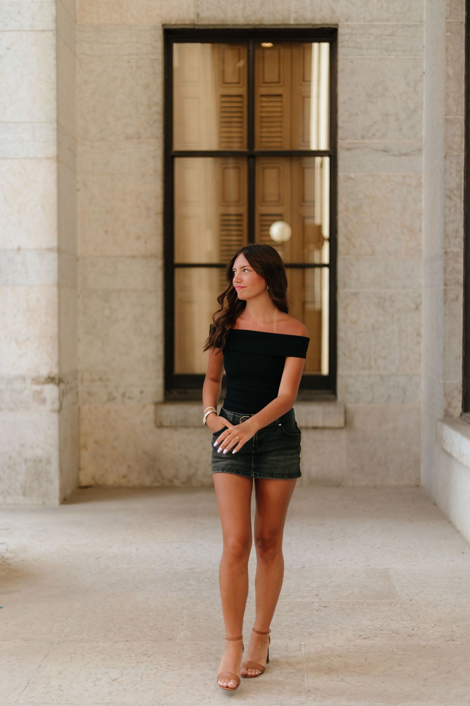 Woman in black dress walks through elegant stone archway corridor with wooden shuttered windows.