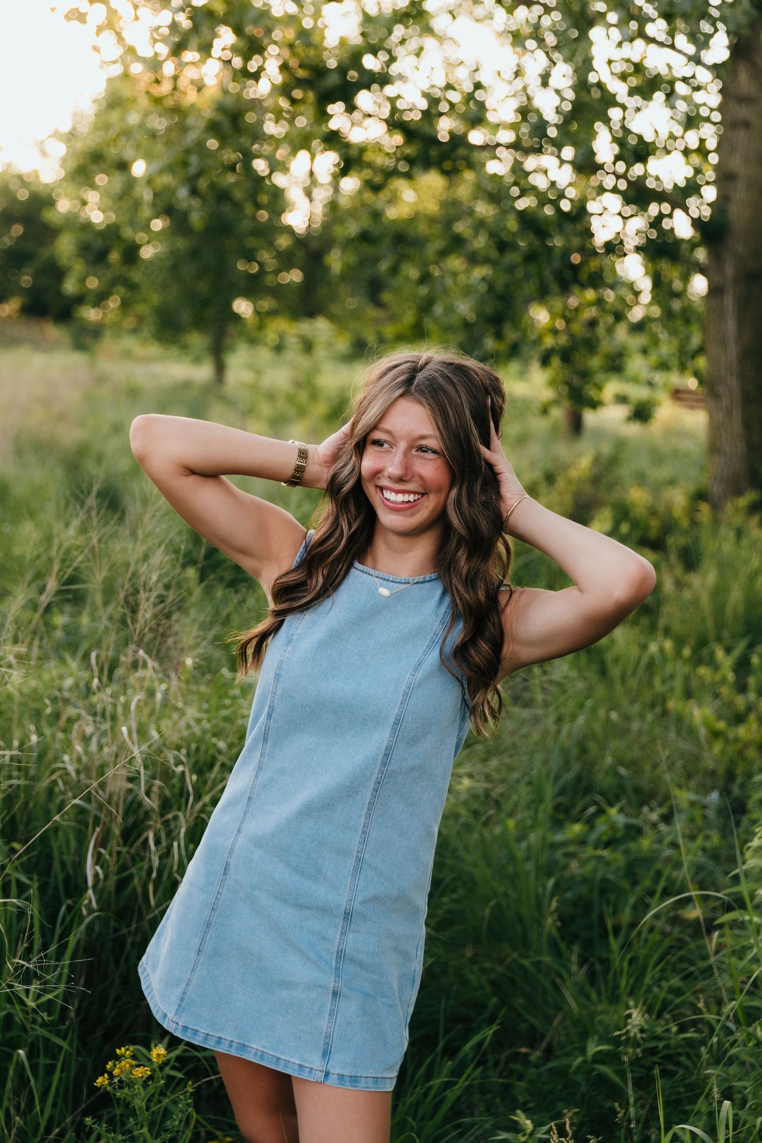 Young woman in denim dress smiling outdoors in lush green field with trees, hands in wavy brown hair.