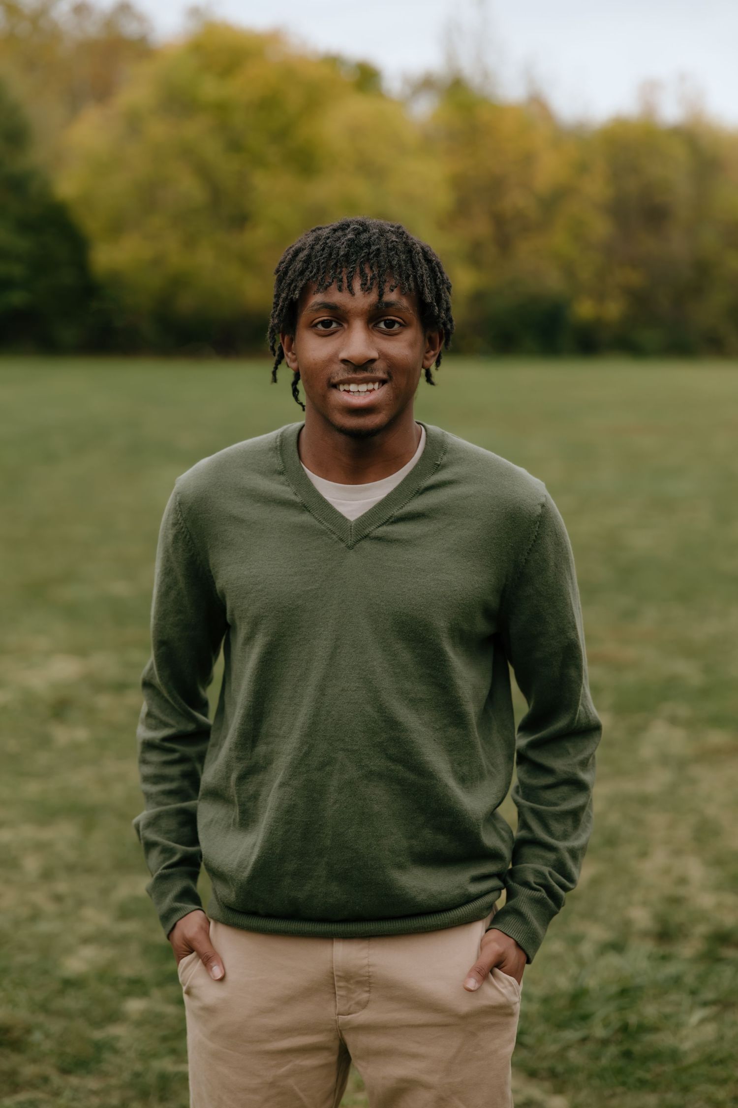 A person wearing a green sweater and khaki pants stands in a grassy field on an autumn day.