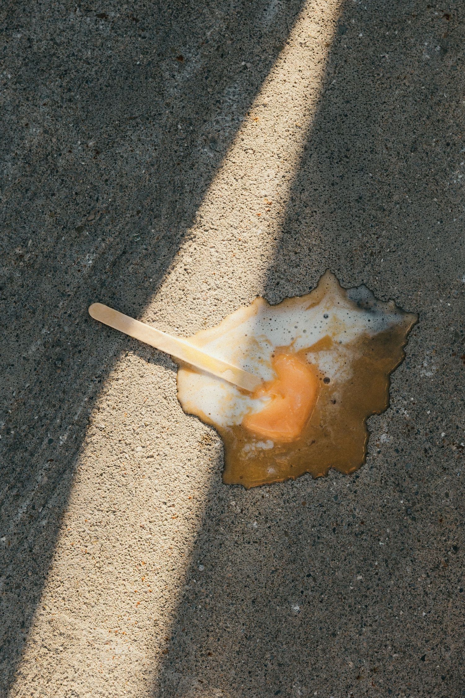Melted orange creamsicle ice cream bar with wooden stick dropped on gray asphalt pavement in sunlight.