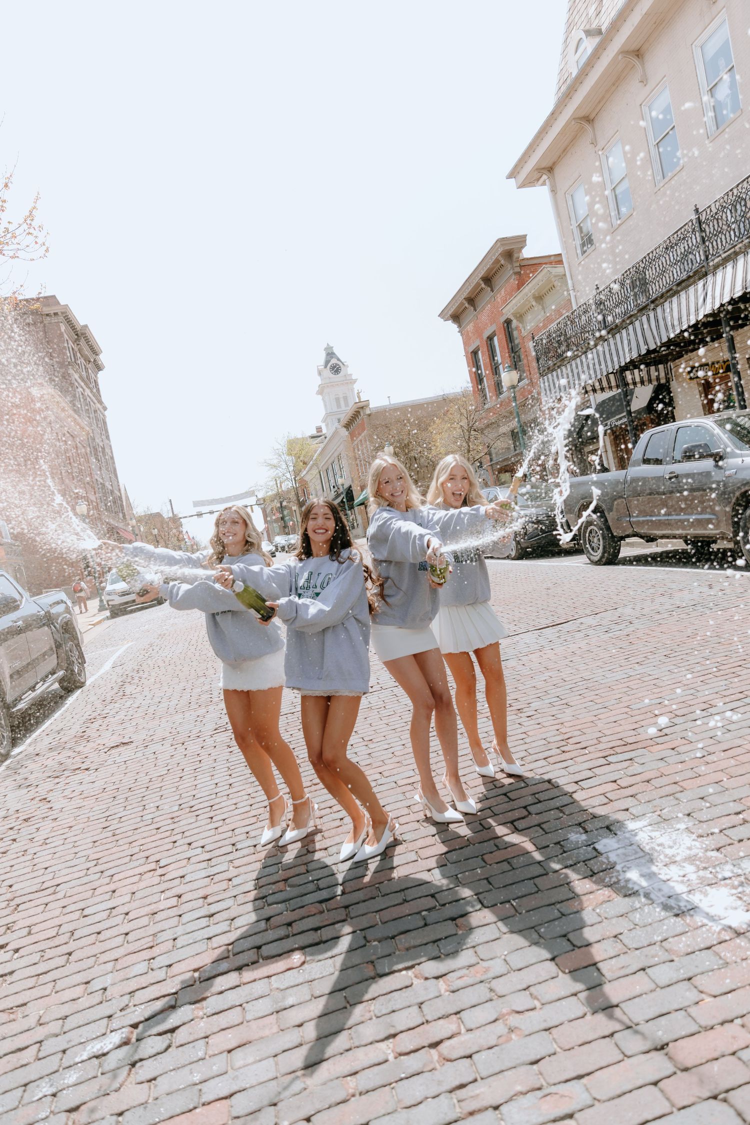 A group in matching gray sweaters jumps together on a cobblestone street while celebrating a joyful moment.