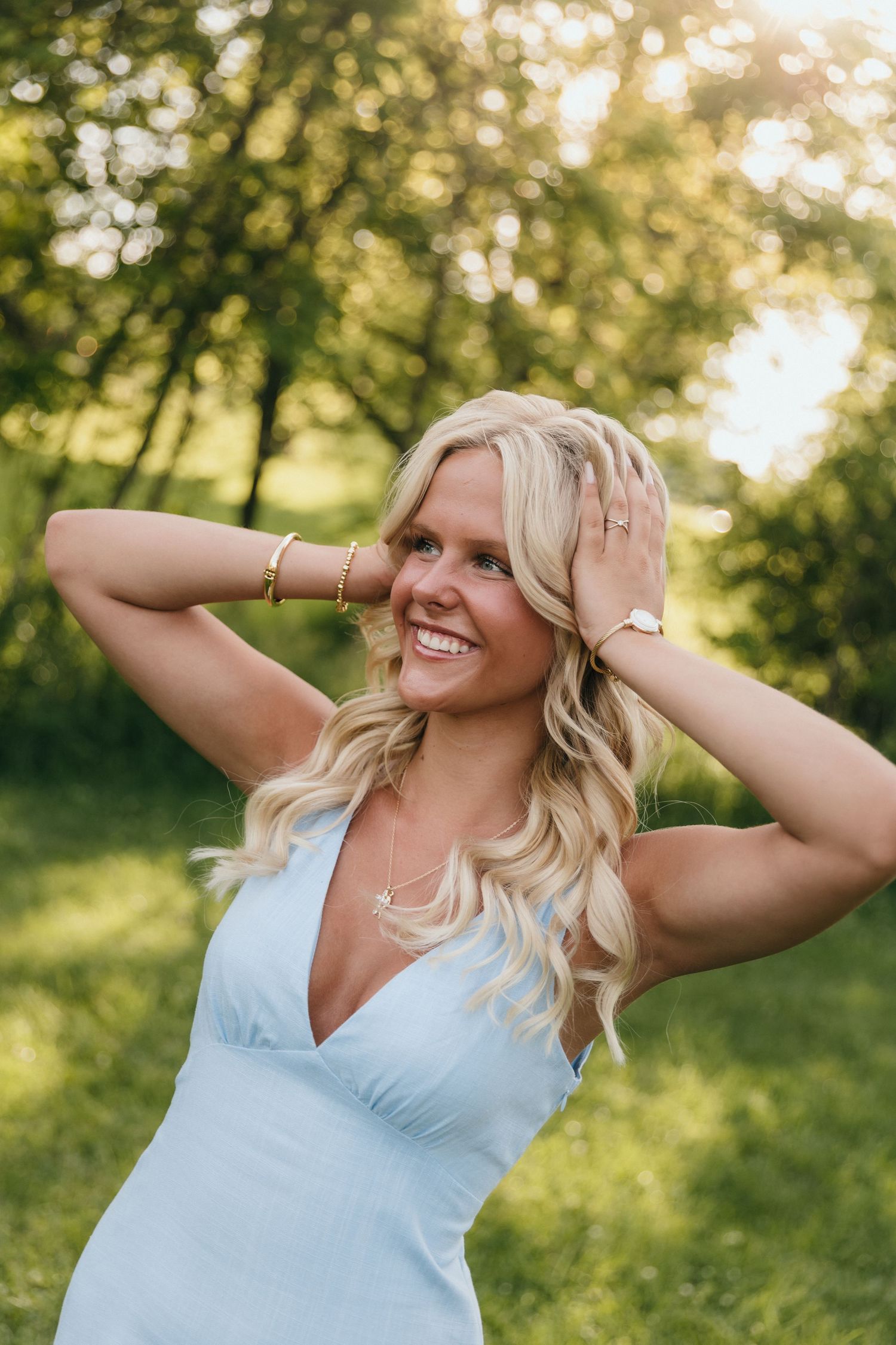 Smiling blonde woman in light blue dress posing outdoors with hands in hair, wearing gold jewelry in sunny park.