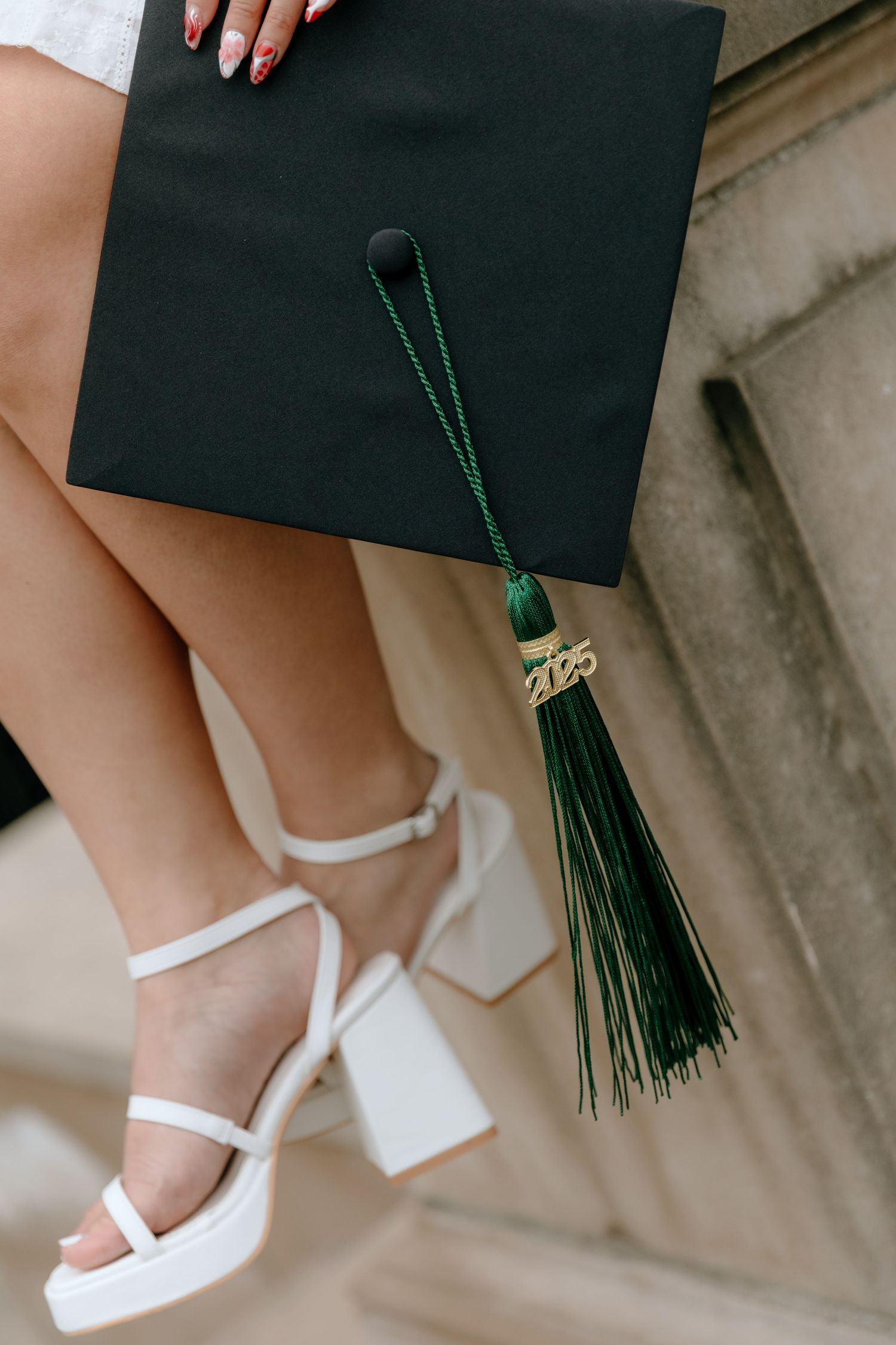 Graduate wearing black graduation gown and white platform sandals poses with green tassel on stairs.