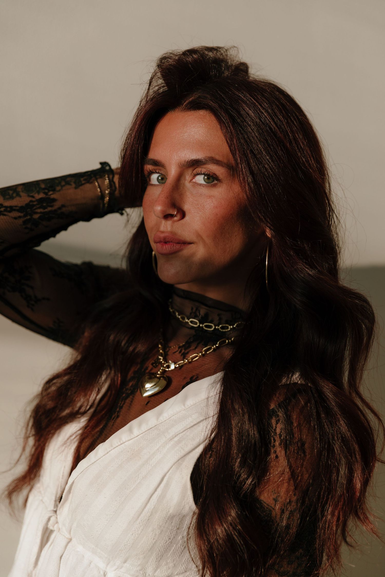 Portrait of person with long dark hair wearing white top and layered necklaces in moody lighting against neutral background.