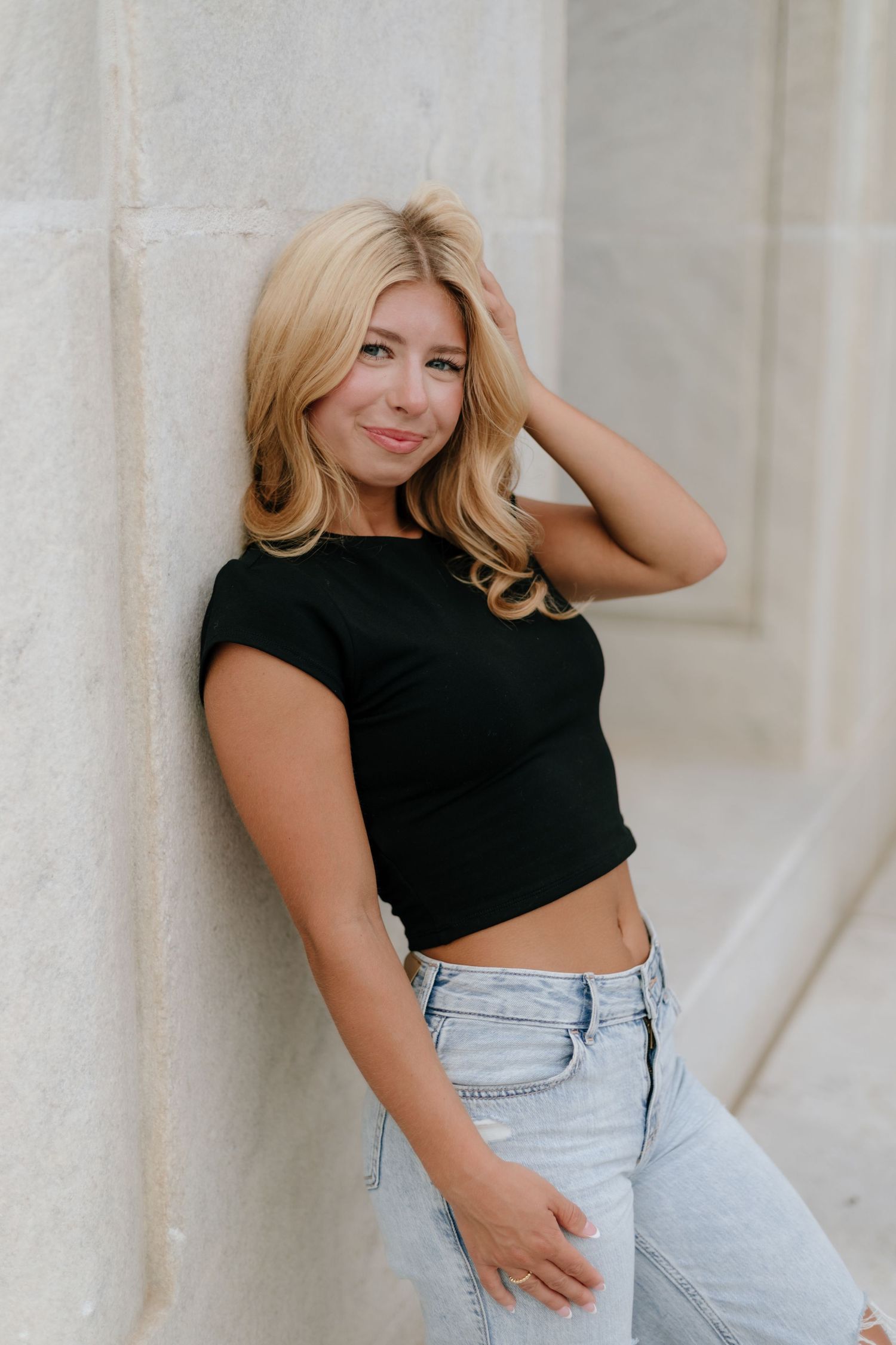 Young woman in black crop top and jeans poses against white stone wall.
