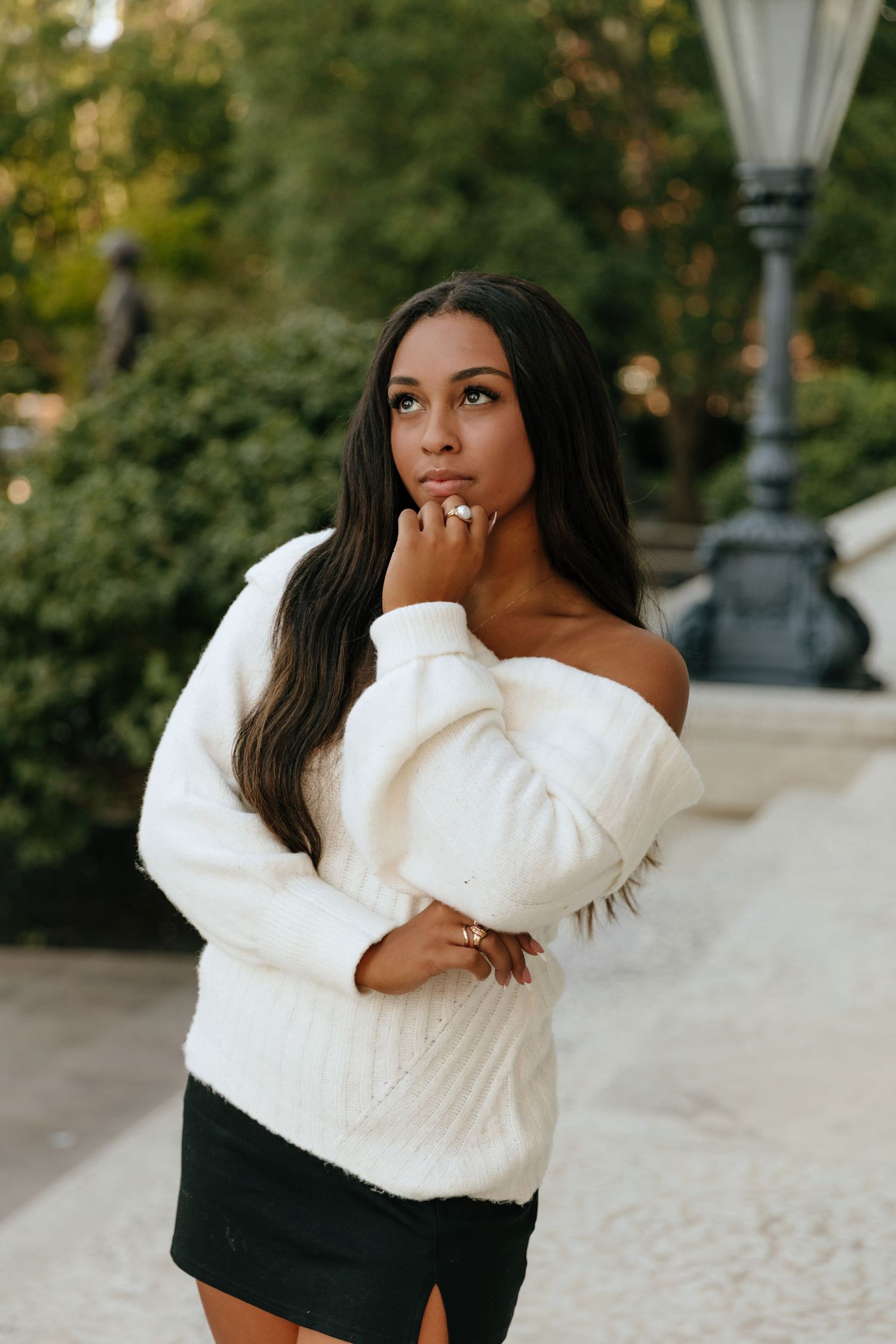 A woman in a cozy white sweater and black pants poses thoughtfully against an outdoor backdrop with lamp posts.