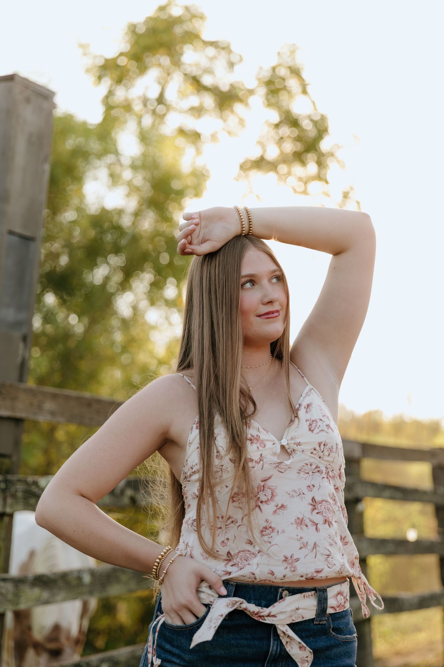 Person in floral tank top and jeans poses casually by wooden fence at sunset in rural setting.