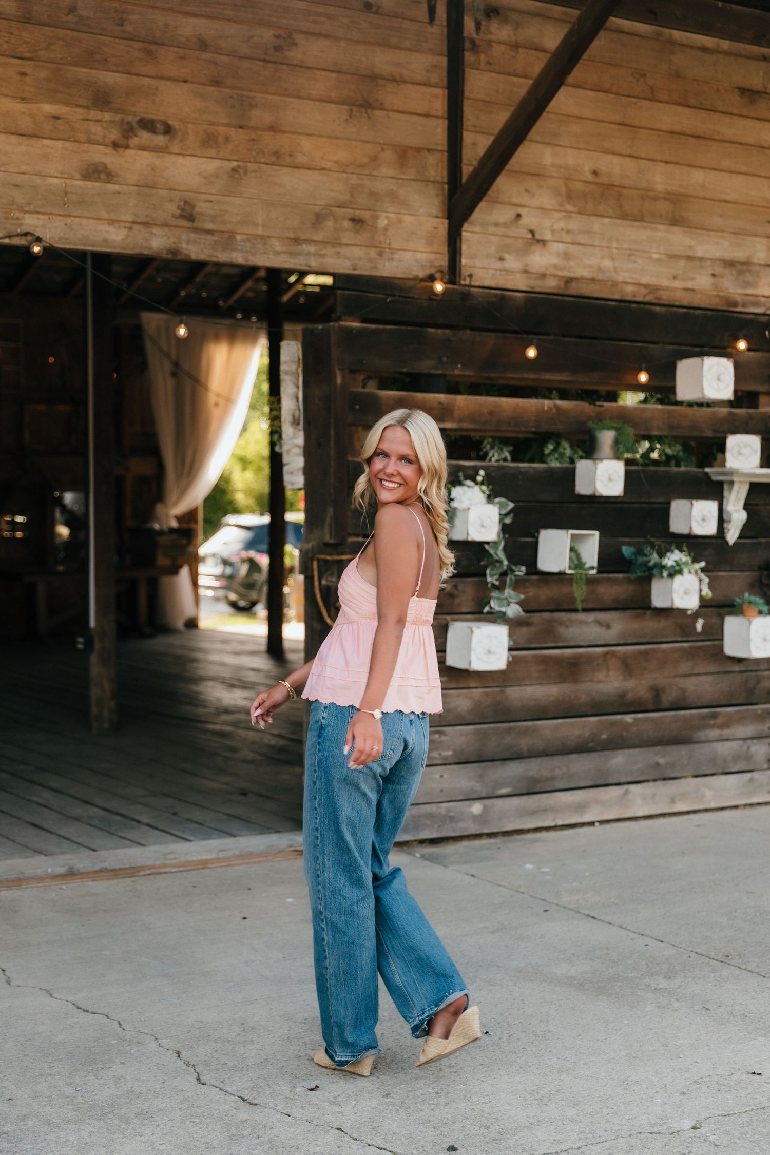 Smiling blonde woman in pink cami top and wide-leg jeans posing outside a rustic wooden barn venue.