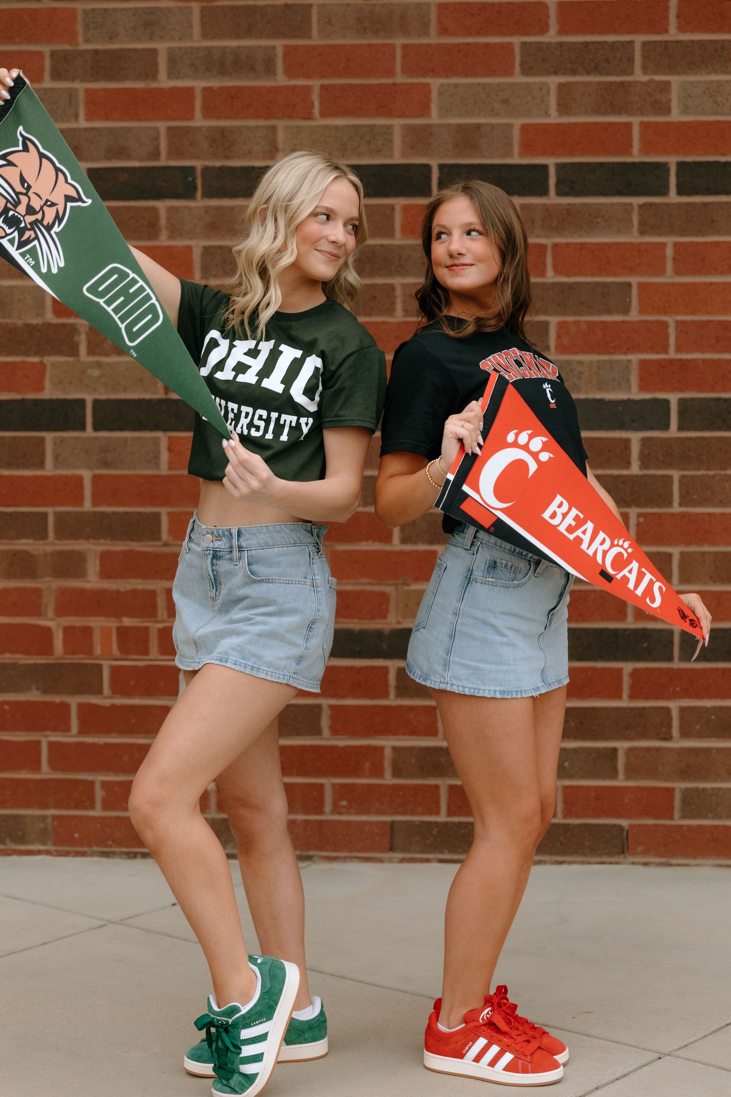 Two people wearing university apparel and denim shorts show off contrasting green and red sneakers against a brick wall.