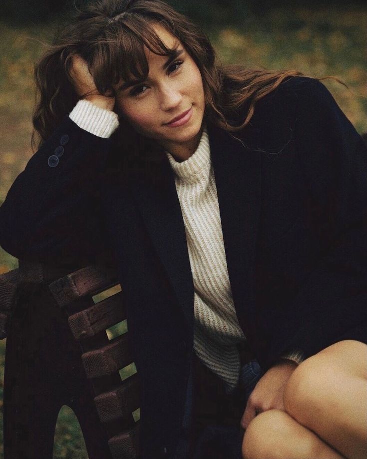 Young woman with wavy brown hair and bangs rests her head on her hand while sitting on a wooden park bench in a navy coat and cream sweater.
