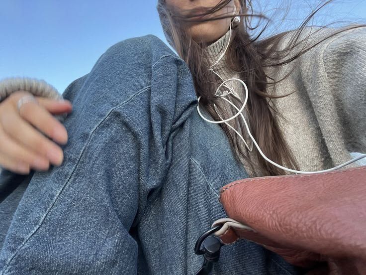 Close-up selfie of a person with long windblown hair wearing a gray knit sweater, denim jacket and white earbuds against a clear blue sky.