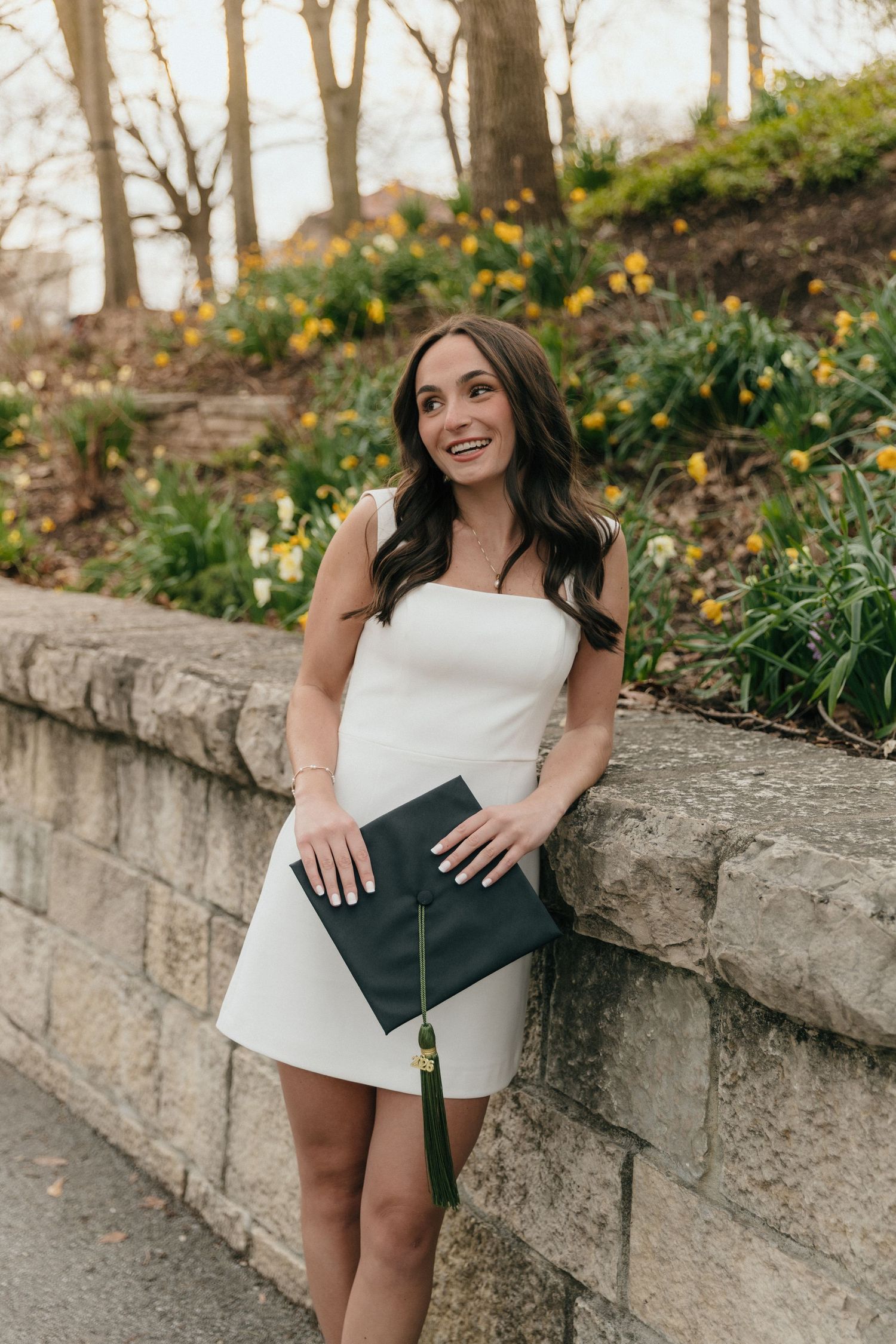 Smiling graduate in white strapless dress holds green mortarboard cap beside spring flower garden wall.