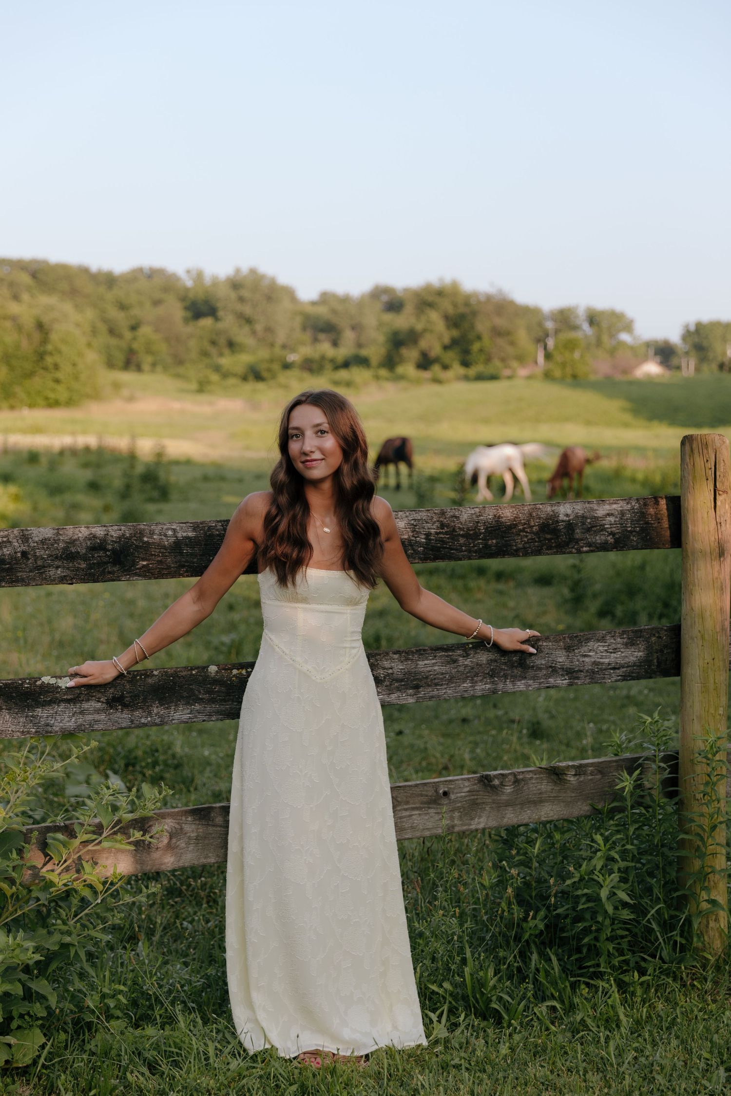 Woman in white dress posing playfully by rustic fence in countryside setting.
