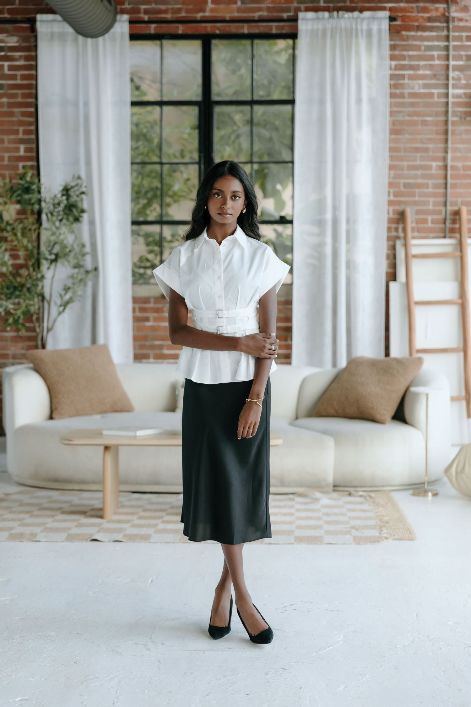 A person stands in a modern living room wearing a white button-down blouse and dark midi skirt with classic black pumps.